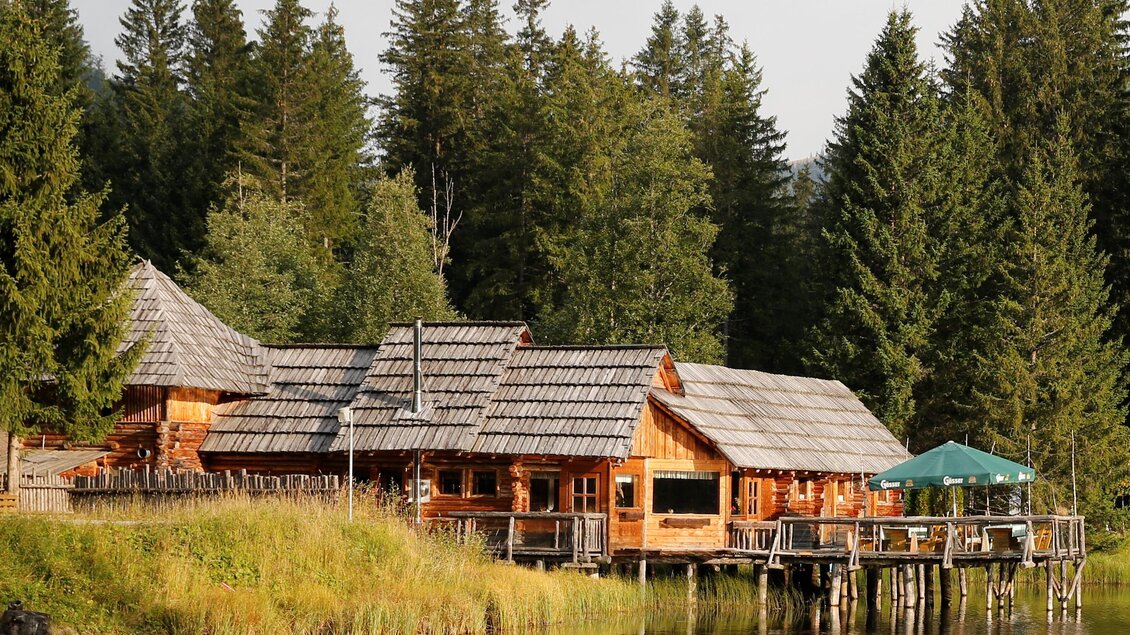 Eine gemütliche Holzferienhütte am Ufer eines ruhigen Sees, umgeben von dichten Wäldern. Über der Hütte schimmert der Himmel in sanften Farben. | © Erlebnisregion Murtal