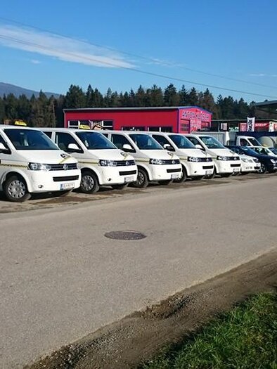 A row of white taxis is parked at a gas station. In the background, trees and a blue sky can be seen. | © Edegger Taxi