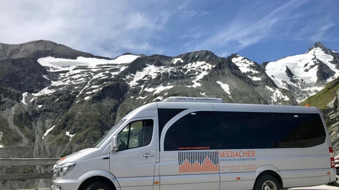 Ein moderner Reisebus parkt an einer Bergstraße mit schneebedeckten Gipfeln im Hintergrund. Der Himmel ist klar und blau, was eine schöne alpine Landschaft ergibt. | © Taxi Seebacher