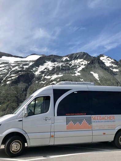 A modern coach is parked along a mountain road with snow-capped peaks in the background. The sky is clear and blue, creating a beautiful alpine landscape. | © Taxi Seebacher
