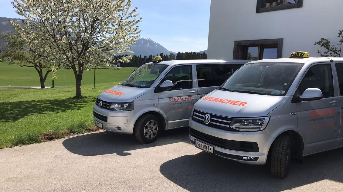 Zwei silberne Taxis stehen auf einem gepflasterten Platz vor einem Gebäude. Im Hintergrund sind Bäume und eine bergige Landschaft sichtbar. | © Taxi Seebacher