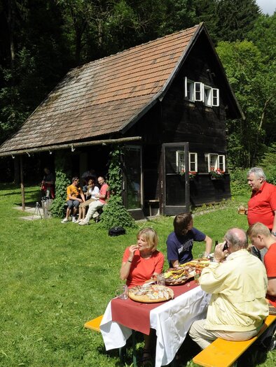 Eine Gruppe von Menschen sitzt an einem Tisch im Freien und genießt ein Picknick. Im Hintergrund steht eine Holzhütte umgeben von Bäumen und Wiese.