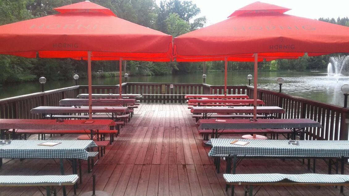 Eine Terrasse am Wasser mit roten Sonnenschirmen und langen Tischen. Die Umgebung ist grün und ruhig, ideal für entspannte Stunden im Freien. | © Muhri & Platz OG;