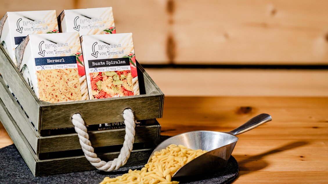 Ein Holzkorb mit verschiedenen Sorten von Pasta in attraktiven Verpackungen steht auf einem Tisch. Daneben liegt eine Metallkelle mit frischer Pasta. | © Region Graz - Mias Photoart