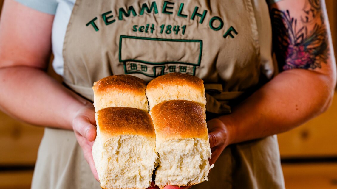 Eine Person hält zwei frische Brötchen in den Händen. Sie trägt eine Schürze mit dem Aufdruck „Temmelhof seit 1847“. | © Region Graz - Mias Photoart