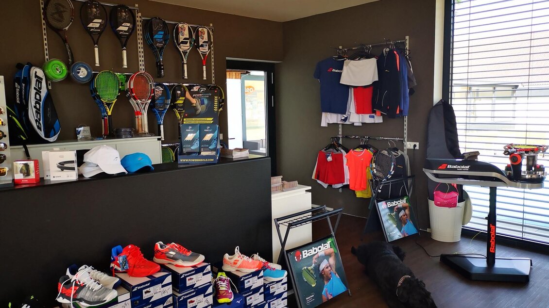 Ein Sportgeschäft mit verschiedenen Sportartikeln wie Schlägern, Kleidung und Schuhen. Im Vordergrund liegen mehrere Schuhkartons, und ein Hund ruht auf dem Boden. | © Kerim Sijercic