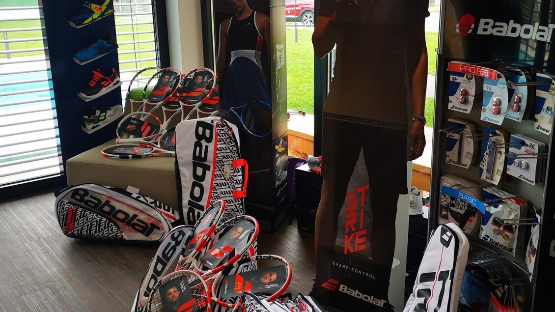 Ein Verkaufsbereich für Tennisartikel mit mehreren Tennisschlägern und einer lebensechten Figur eines Tennisspielers. Im Hintergrund sind Plakate mit Marken wie Babolat zu sehen. | © Kerim Sijercic
