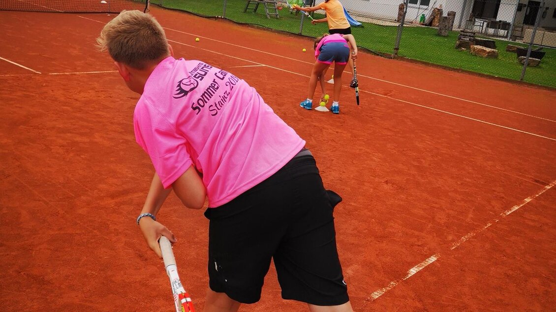 Ein Junge in einem pinken Shirt steht auf einem Tennisplatz und bereitet sich auf einen Schlag vor. Im Hintergrund spielt ein Mädchen Tennis, während ein Haus zu sehen ist. | © Kerim Sijercic