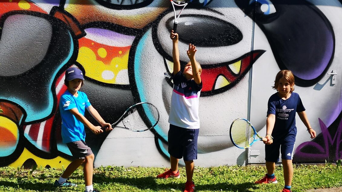 Drei Kinder spielen Tennis vor einer bunten Graffiti-Wand. Sie halten Schläger und freuen sich beim Spiel. | © Kerim Sijercic