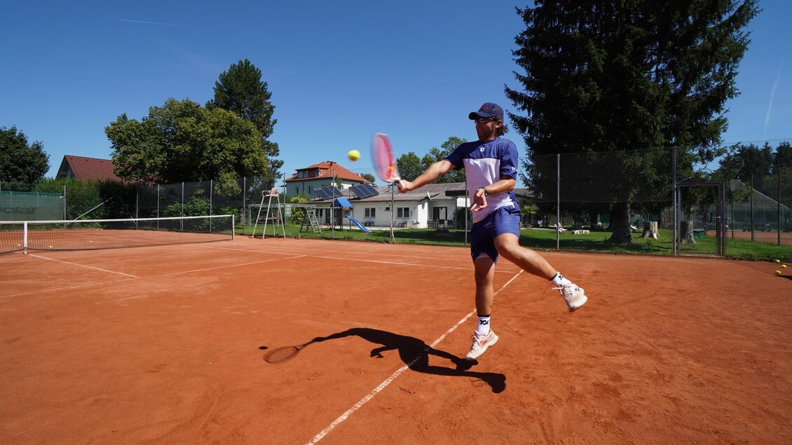 Eine Person spielt Tennis auf einem roten Sandplatz. Im Hintergrund sind Bäume und ein Gebäude sichtbar. | © Kerim Sijercic