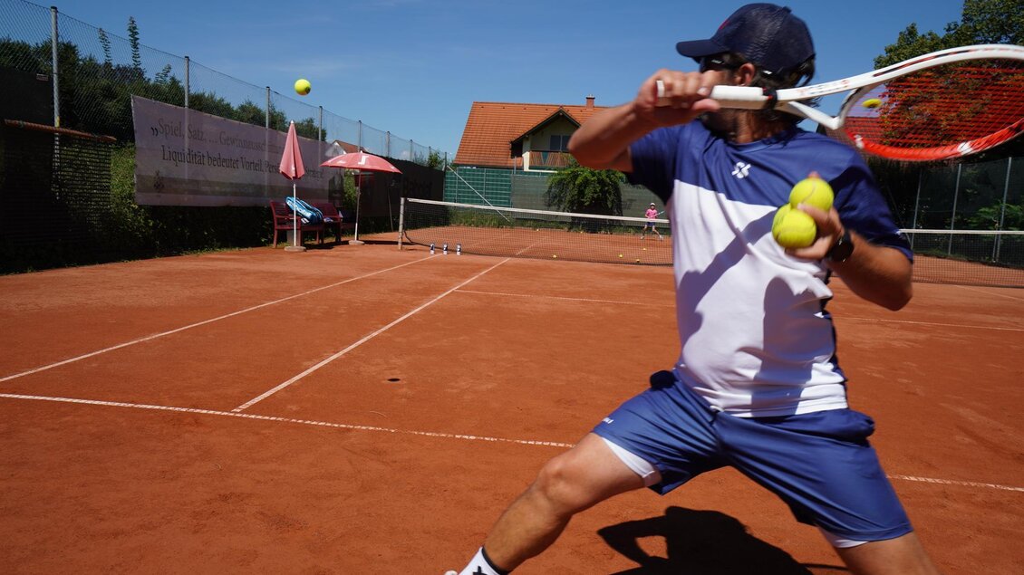 Ein Tennisspieler schlägt einen Ball auf einem roten Platz. Er hält mehrere Tennisbälle in der Hand und steht unter klarem Himmel. | © Kerim Sijercic