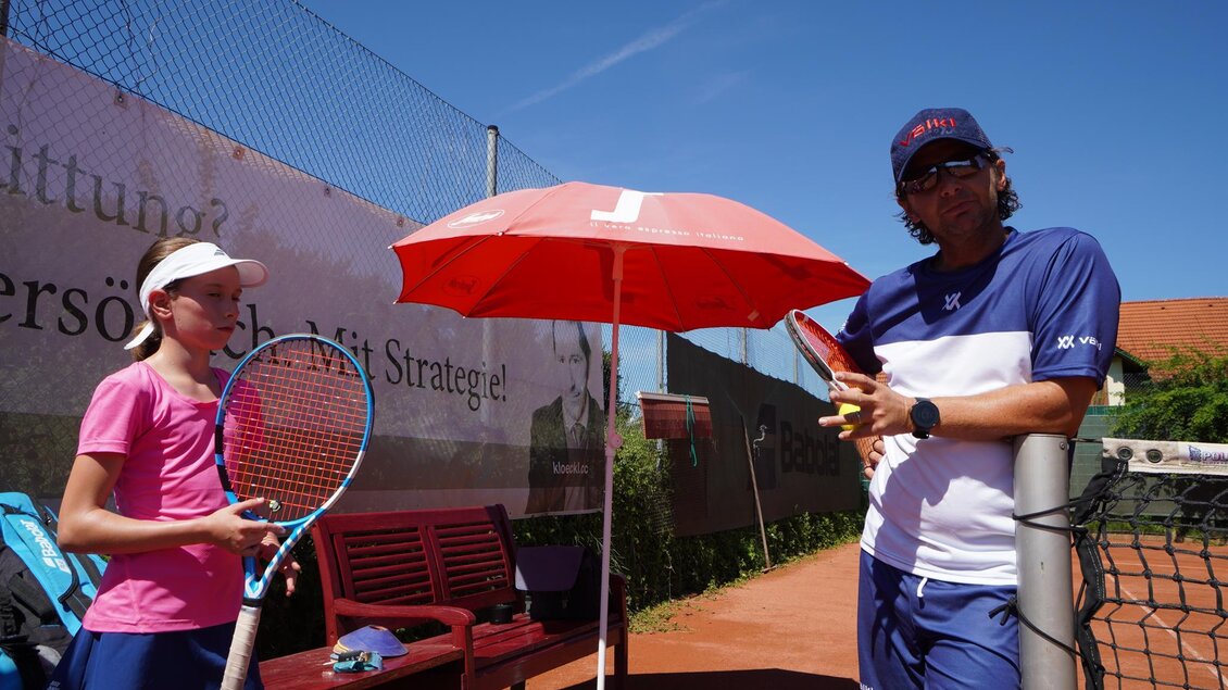 Ein Tennisplatz mit einem Trainer und einem jungen Spieler. Beide stehen unter einem Sonnenschirm und haben Tennisschläger in der Hand. | © Kerim Sijercic