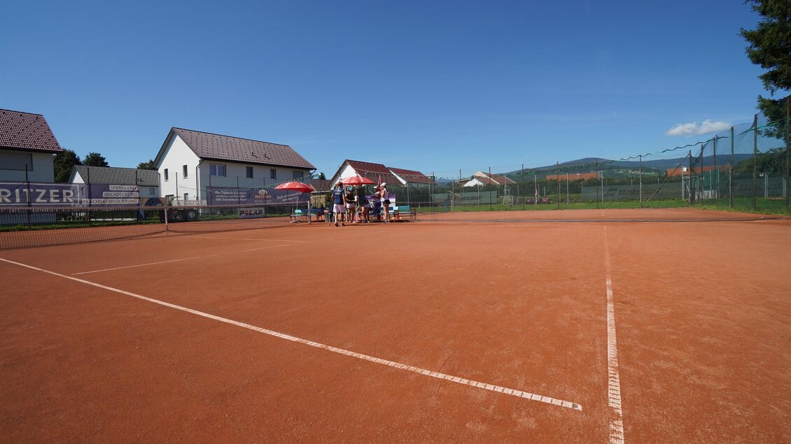 Ein Tennisplatz aus rotem Sand unter einem klaren blauen Himmel. Im Hintergrund sind einige Wohnhäuser und ein Tennisnetz zu sehen. | © Kerim Sijercic