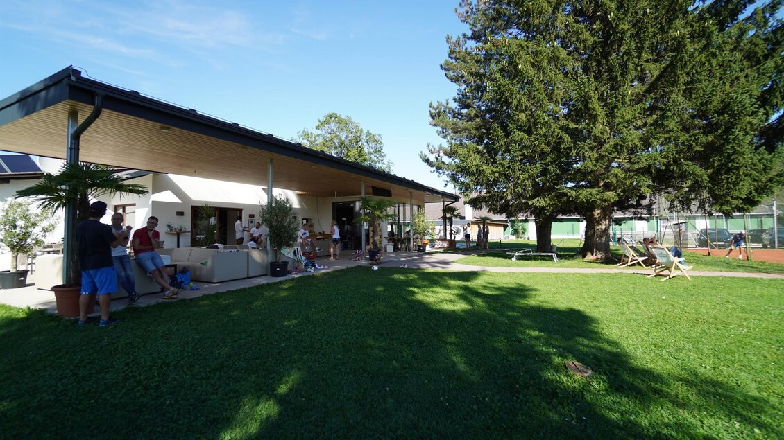 Ein modernes Gebäude mit einer großen Rasenfläche und schattenspendenden Bäumen. Menschen entspannen und unterhalten sich im Freien. | © Kerim Sijercic