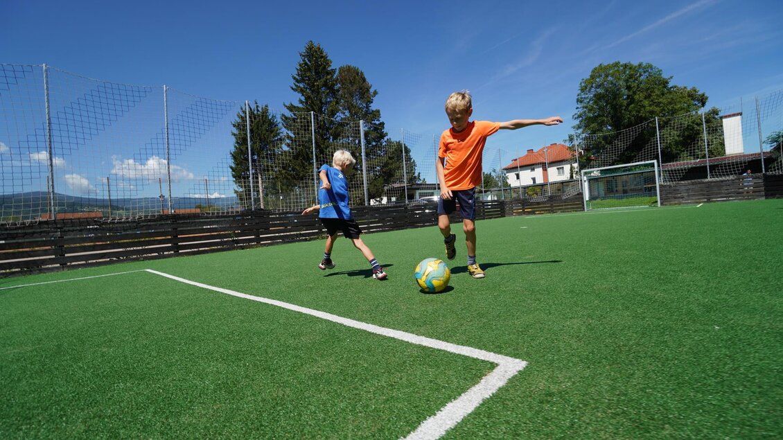 Zwei Kinder spielen auf einem Fußballplatz aus Kunstrasen. Ein Junge dribbelt den Ball, während der andere zuschaut. | © Kerim Sijercic