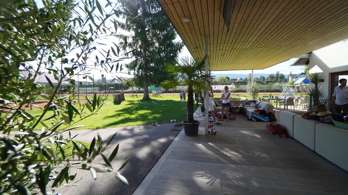 Ein sonniger Außenbereich mit einer Holzterrasse und Pflanzen. Im Hintergrund sind Menschen zu sehen, die sich auf einer grünen Wiese aufhalten. | © Kerim Sijercic