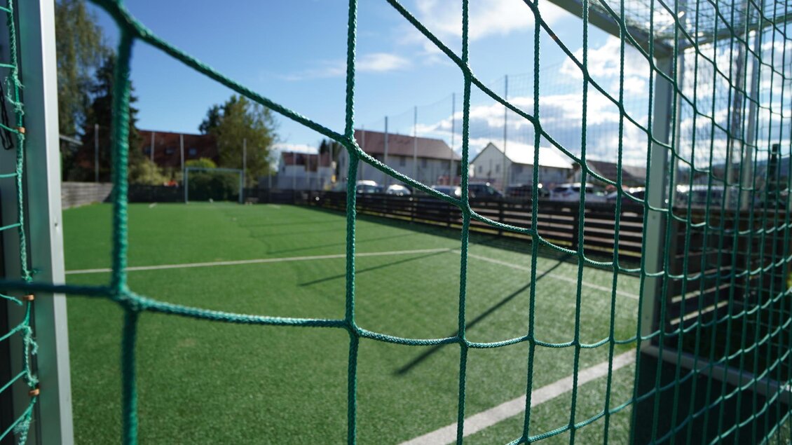 Ein Fußballplatz mit Kunstrasen und einem klaren blauen Himmel. Im Hintergrund sind Häuser und Bäume sichtbar. | © Kerim Sijercic