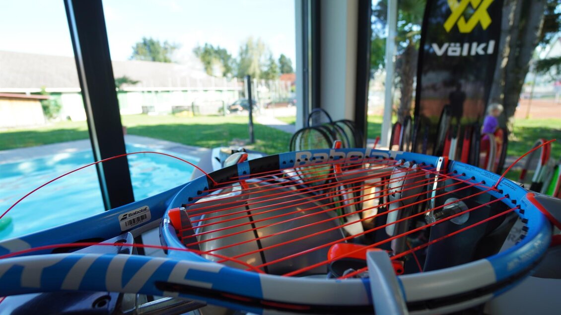 Ein Tennisracket mit rotem Saiten ist in einem Raum positioniert, der einen Blick auf einen Pool im Freien bietet. Im Hintergrund sind einige Tennisartikel und Bäume zu sehen. | © Kerim Sijercic