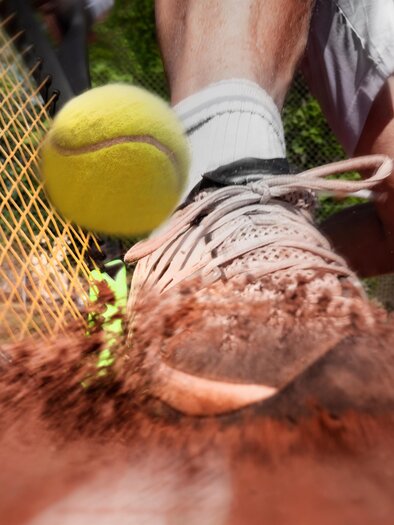 A tennis ball bounces off the ground while a player with a tennis racket is active in the background. The scene captures a dynamic moment on a red clay court. | © AdobeStock