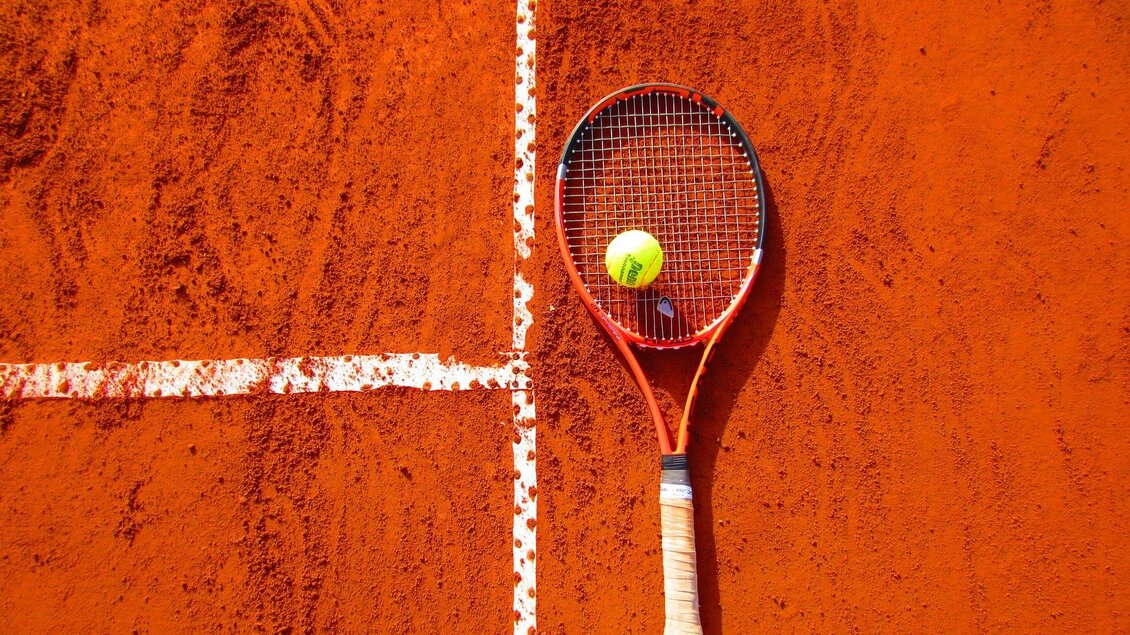 Ein Tennisplatz mit rotem Sand und weißen Linien. Ein Tennisschläger liegt neben einem gelben Tennisball. | © Pixabay/Cynthiamcastro