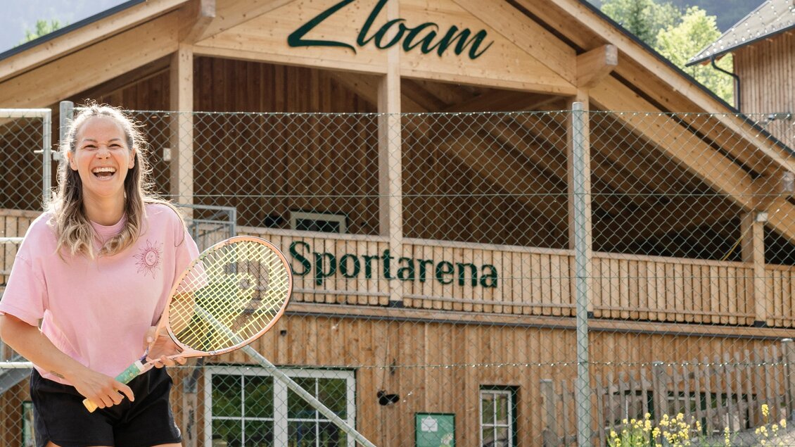 Eine fröhliche junge Frau hält einen Tennisschläger vor einem Sportarena mit Holzbau. Im Hintergrund ist der Schriftzug "Zloam" zu sehen. | © www.zloam.at