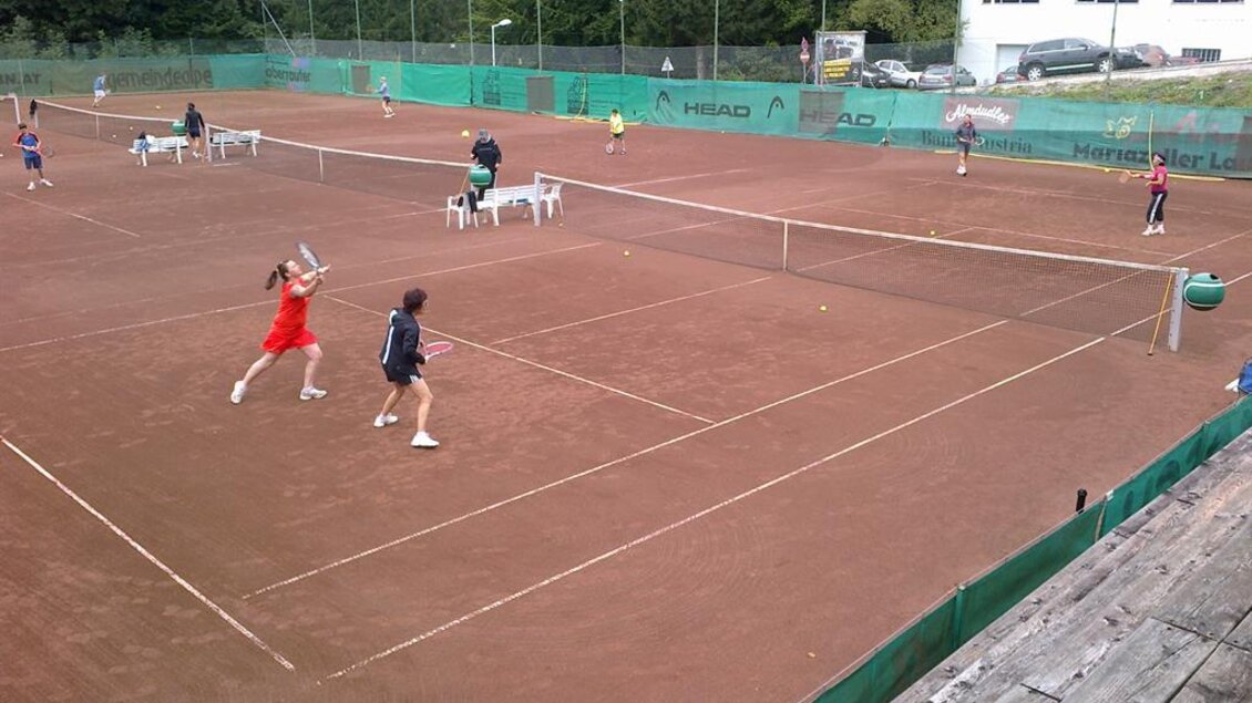 Ein Tennisplatz mit mehreren Spielern in Aktion. Im Hintergrund sind Bäume und ein großes Gebäude sichtbar. | © UTC Mariazell