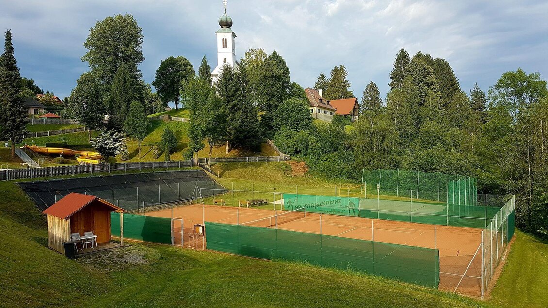 Ein Tennisplatz umgeben von grünen Wiesen und Bäumen. Im Hintergrund ist ein Kirchturm zu sehen. | © Tennisplatz St. Oswald ob Eibiswald