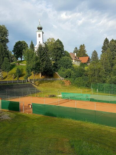 A tennis court surrounded by green meadows and trees. In the background, a church tower can be seen. | © Tennisplatz St. Oswald ob Eibiswald