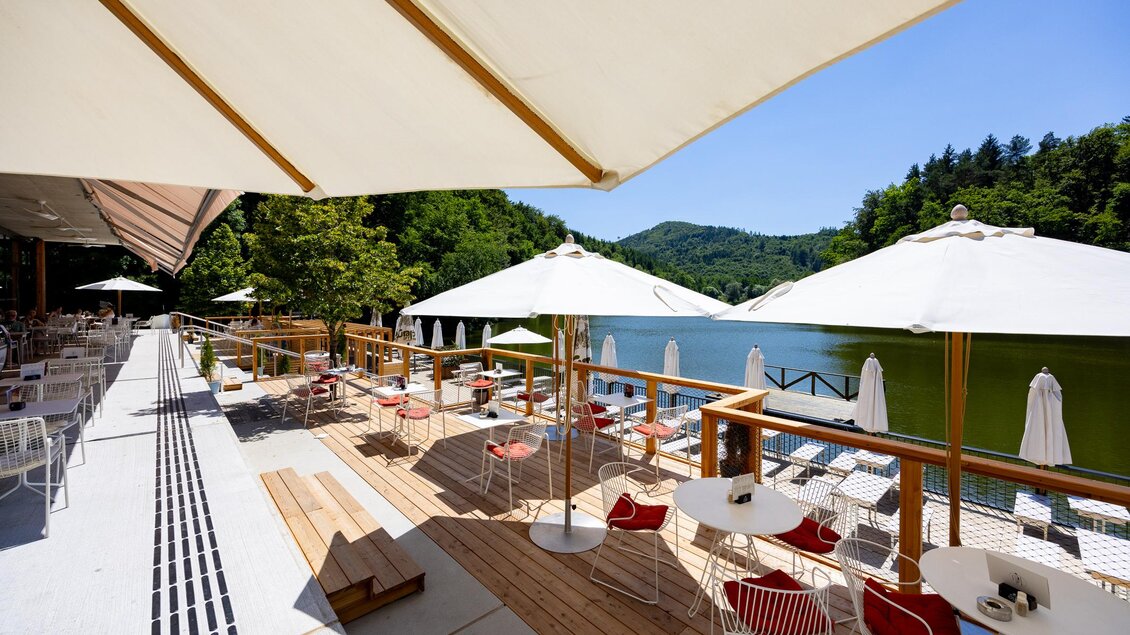 Ein gemütlicher Außenbereich mit Tischen und Stühlen am Wasser. Helle Sonnenschirme spenden Schatten vor einer malerischen Kulisse. | © Region Graz, Harry Schiffer