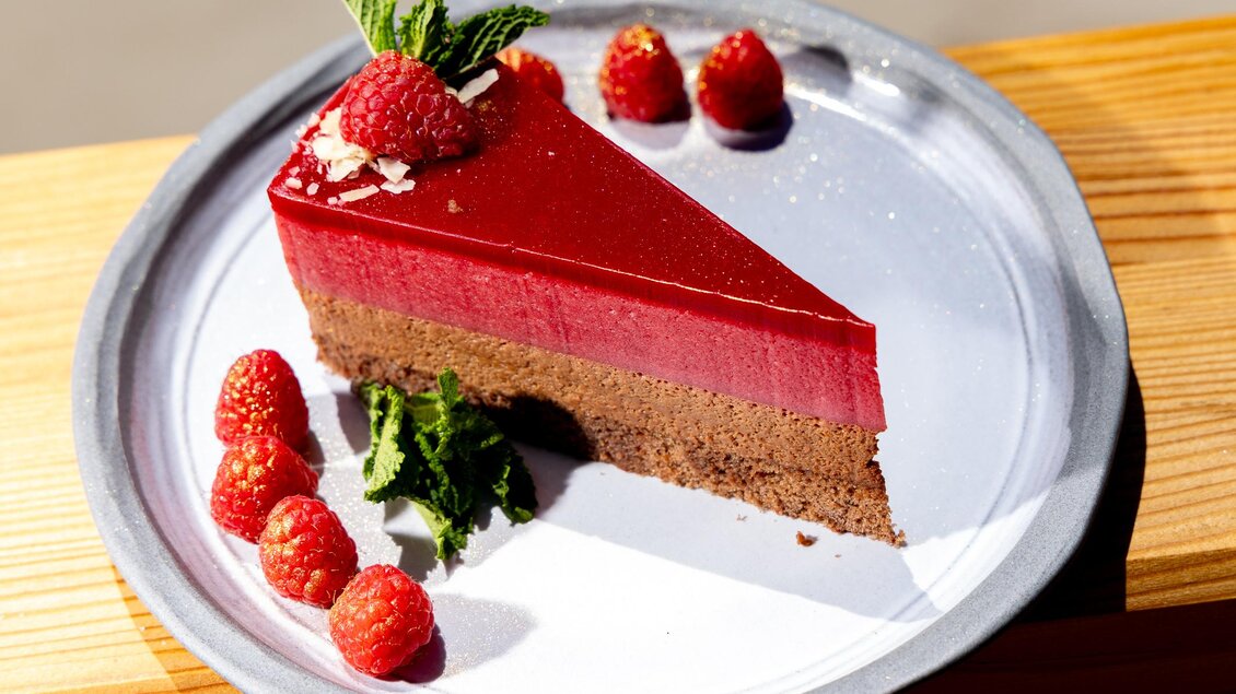 Ein Stück Himbeertorte auf einem Teller, dekoriert mit frischen HimBeeren und Minze. Die Torte hat eine schokoladige Basis und eine glänzende Fruchtschicht. | © Region Graz, Harry Schiffer