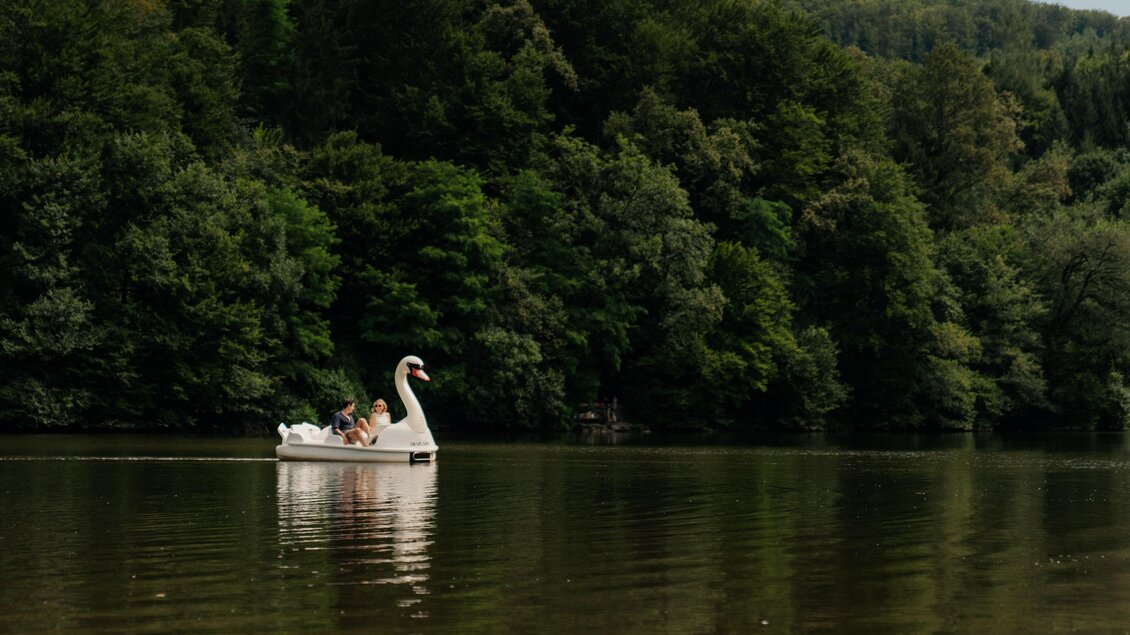 Ein weißes Schwanenboot schwimmt ruhig auf einem See. Im Hintergrund sind dicht bewaldete Hügel zu sehen. | © (c) Region Graz - studio draussen