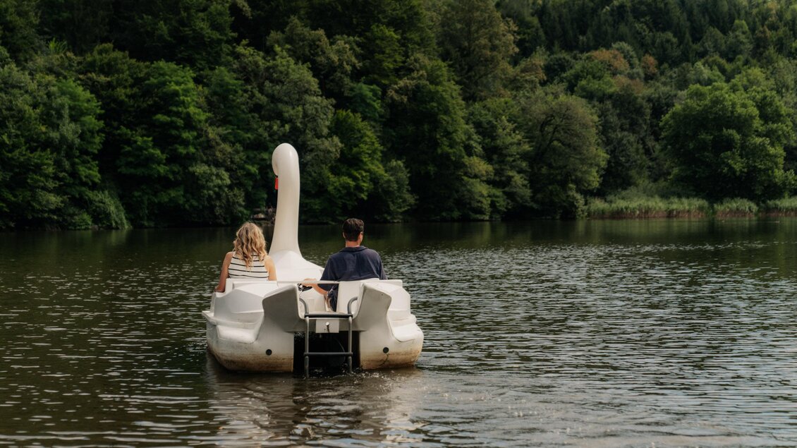 Ein Paar fährt auf einem Schwanschnabelboot über einen ruhigen See. Umgeben von grünen Bäumen genießt es die Natur. | © (c) Region Graz - studio draussen