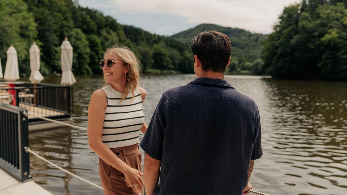 Ein Paar steht am Ufer eines ruhigen Sees, umgeben von üppigem Grün. Die Frau trägt eine gestreifte Bluse und Sonnenbrille, während der Mann sie ansieht. | © (c) Region Graz - studio draussen