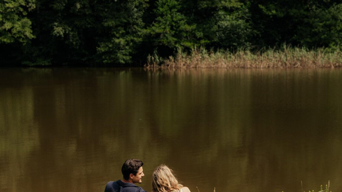 Ein Paar sitzt auf einer Bank am Ufer eines stillen Gewässers. Umgeben von grünen Bäumen genießen sie die Natur und die Ruhe. | © (c) Region Graz - studio draussen