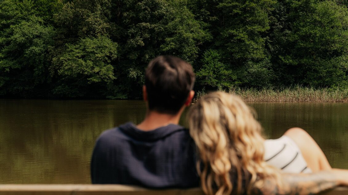 Ein Paar sitzt auf einer Bank und schaut auf einen ruhigen See. Umgeben von grünen Bäumen strahlt die Szene Ruhe und Zweisamkeit aus. | © (c) Region Graz - studio draussen
