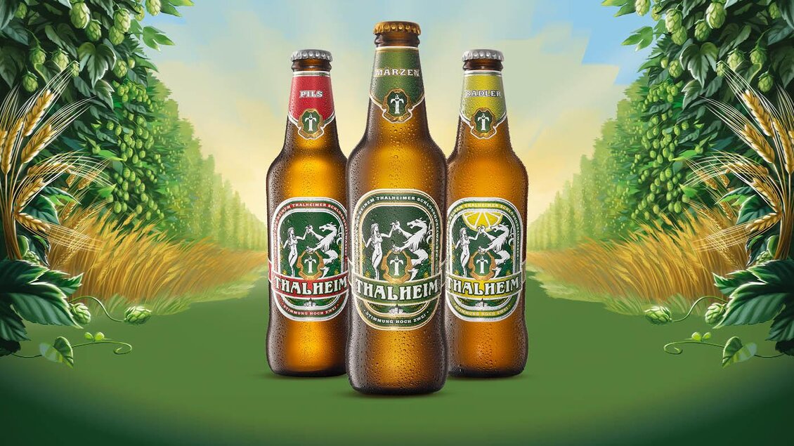 Drei Flaschen Bier stehen in der Mitte, umgeben von grünen Pflanzen. Im Hintergrund erstreckt sich eine sonnige Landschaft mit Weizenfeldern. | © Thalheimer Heilwasser GmbH Brauerei