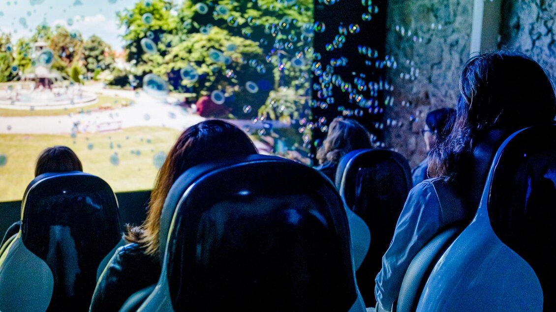 Eine moderne Installation mit Besucher*innen, die in attraktiven Sesseln sitzen. Im Hintergrund ist eine Projektion mit Naturmotiven und Seifenblasen sichtbar. | © Region Graz - Mias Photoart