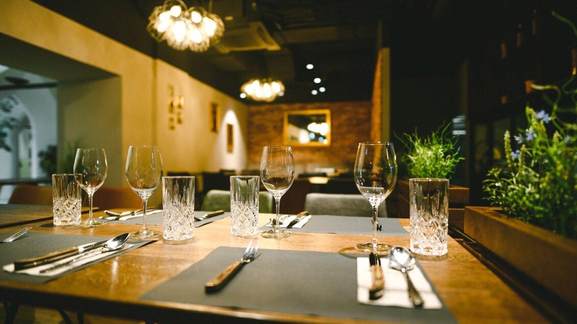 Ein eleganter Tisch in einem Restaurant mit Gläsern und Besteck. Im Hintergrund sind sanfte Lichter und eine stilvolle Inneneinrichtung zu sehen. | © The Kitchen