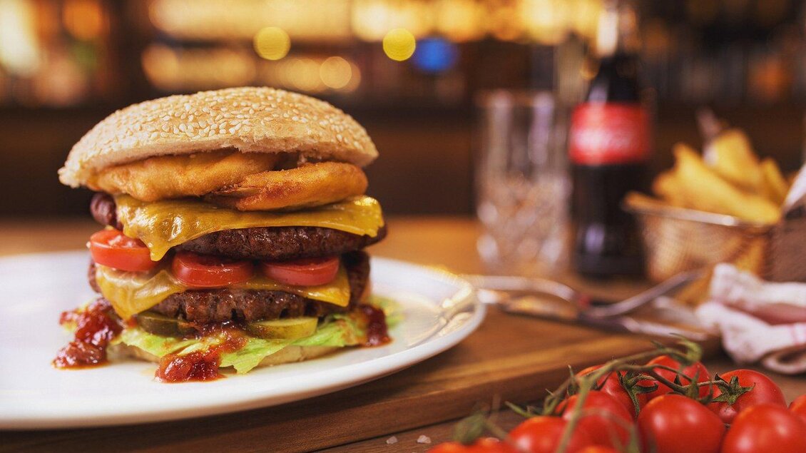Ein saftiger Burger mit Käse, Tomaten und Salat auf einem weißen Teller. Daneben liegen frische Cherrytomaten und eine Flasche Cola. | © The Kitchen