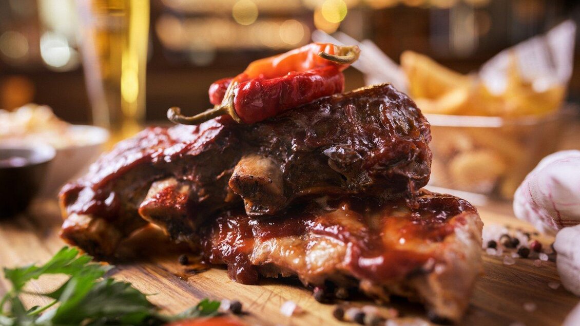 Saftige Rippchen mit würziger Sauce und einer Chilischote obendrauf. Im Hintergrund sind Pommes und ein Glas Bier zu sehen. | © The Kitchen