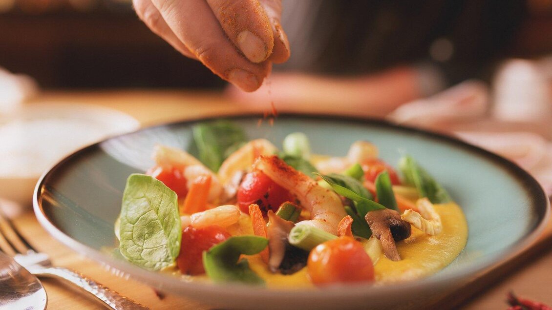 Ein köstlicher Salat mit frischen Zutaten wie Tomaten und Spinat. Eine Hand streut gerade Gewürze über das Gericht. | © The Kitchen