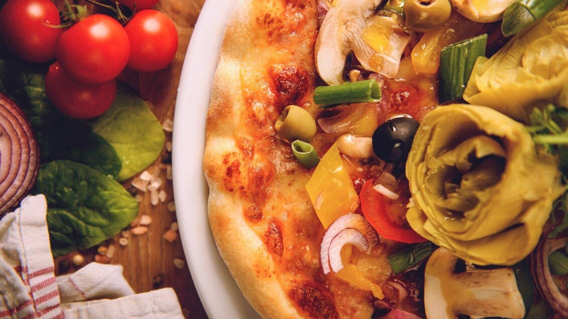 Eine köstliche Pizza mit frischem Gemüse und Käse. Neben der Pizza liegen rote Tomaten und grüne Blattgemüse. | © The Kitchen