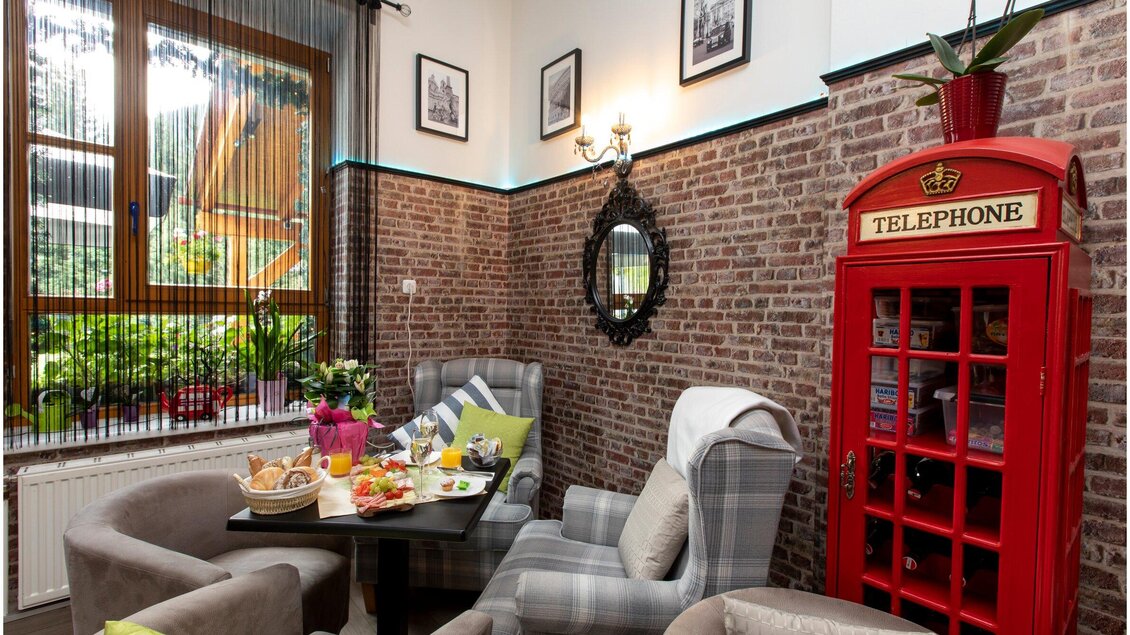 Gemütlicher Innenraum mit Ziegelsteinmauer, stilvollen Sesseln und einem roten Telefonhäuschen. Auf dem Tisch liegen frisches Obst und Snacks, umgeben von Pflanzen und Bildern an der Wand. | © TV Region Graz - Lunghammer