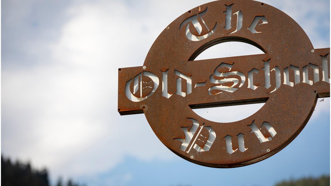 Ein rustikales Schild des "Old-School Pubs" mit stilvollen Buchstaben. Der Hintergrund zeigt einen klaren Himmel und Berge. | © TV Region Graz - Lunghammer