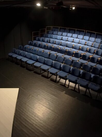An empty auditorium with blue chairs. The lighting is dimmed and the stage is visible. | © THEO Studiobühne