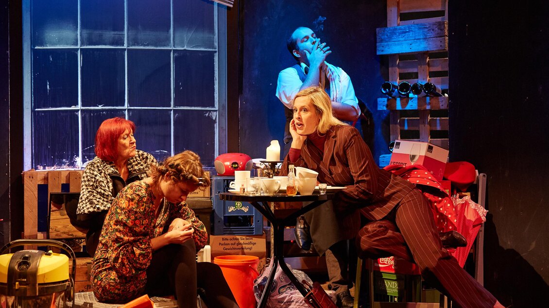 Eine Theateraufführung mit drei Schauspielerinnen und einem Schauspieler. Sie sitzen in einem Raum voller Requisiten und scheinen ein intensives Gespräch zu führen. | © THEO Studiobühne