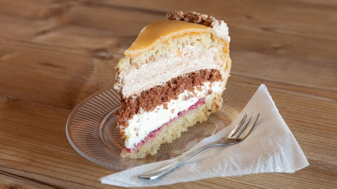 Ein Stück Torte auf einem Teller, das aus verschiedenen Schichten besteht. Die Torte hat eine glatte, gelbe Oberseite und ist von einer Gabel und einer Serviette begleitet. | © TV Region Graz - Harry Schiffer