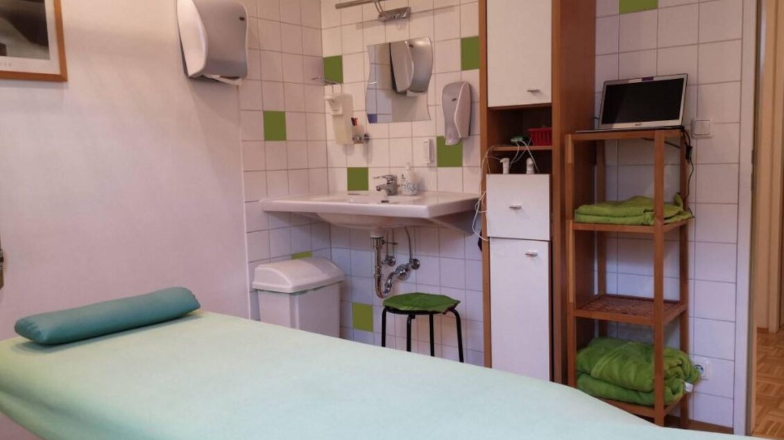 Ein heller Behandlungsraum mit einer Liege und einem Regal. An der Wand befinden sich Handtücher und weitere Pflegeutensilien. | © Therapiezentrum Prä-Fit