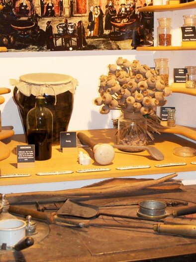 Eine historische Werkstatt mit verschiedenen Gläsern und Utensilien. Auf Regalen sind zahlreiche Kräuter und Gewürze ausgestellt. | © Theriak Museum