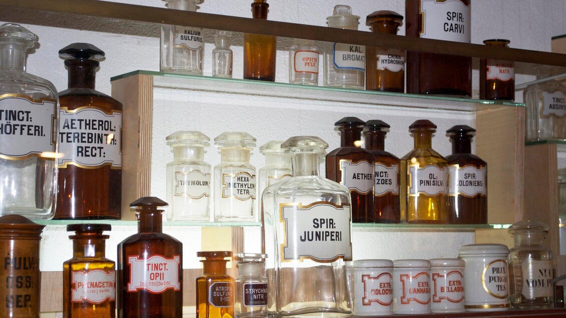 Eine Sammlung von alten Apothekerflaschen auf einem Regal. Die Flaschen sind in verschiedenen Größen und Farben, mit gut lesbaren Etiketten. | © Theriak Museum
